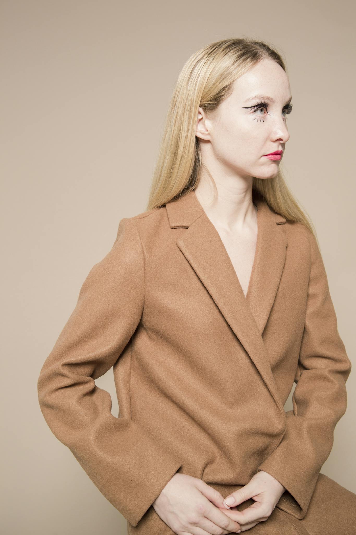 A poised woman in a beige blazer gazes confidently in a minimalistic studio setting.