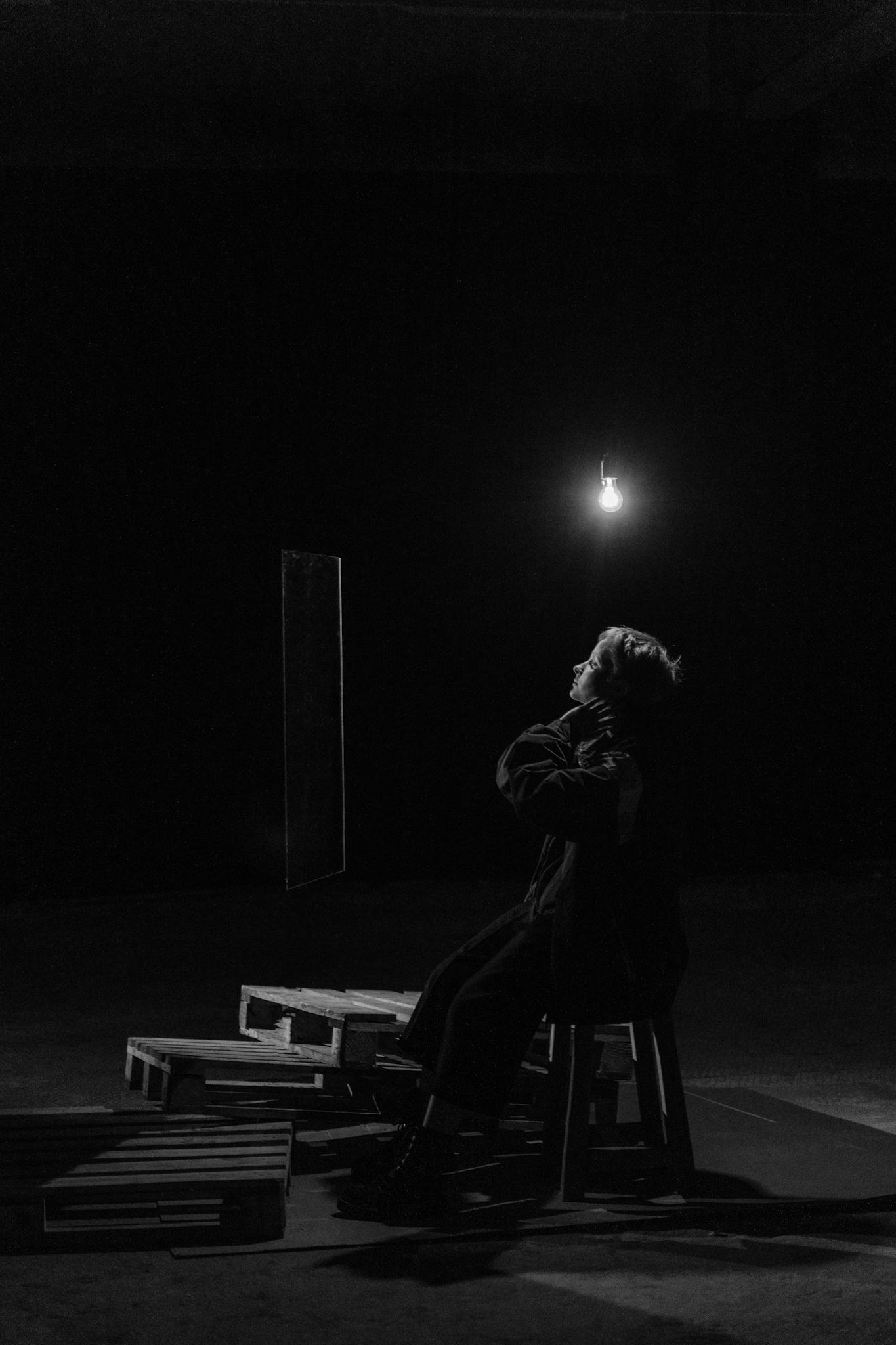 A solitary figure poses thoughtfully in a dark studio, illuminated by a single bulb.